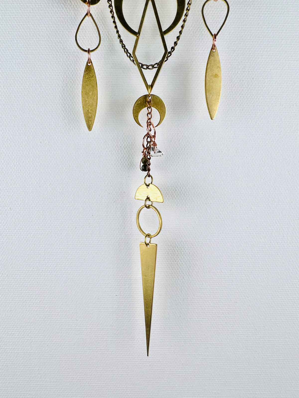 Gold colored metal shapes and wire wrapped Green tourmaline and Herkimer diamond beads connected by small copper rings creating three separate vertical hanging pieces in the bottom portion of a piece of crystal wall art. Shapes include drops, ovals, diamonds, crescent moon, and half moon with a sharp long triangle at the bottom. 