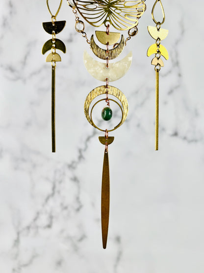 Three vertically hanging sections of connected gold colored metal and cream colored acrylic shapes make up the bottom portion of this hanging crystal wall art. Shapes include, Monstera leaf, teardrop shapes, half circles, crescent moons, long thin gold bars, and a wire wrapped green bead. All hang in front of a gray and white marble wall.