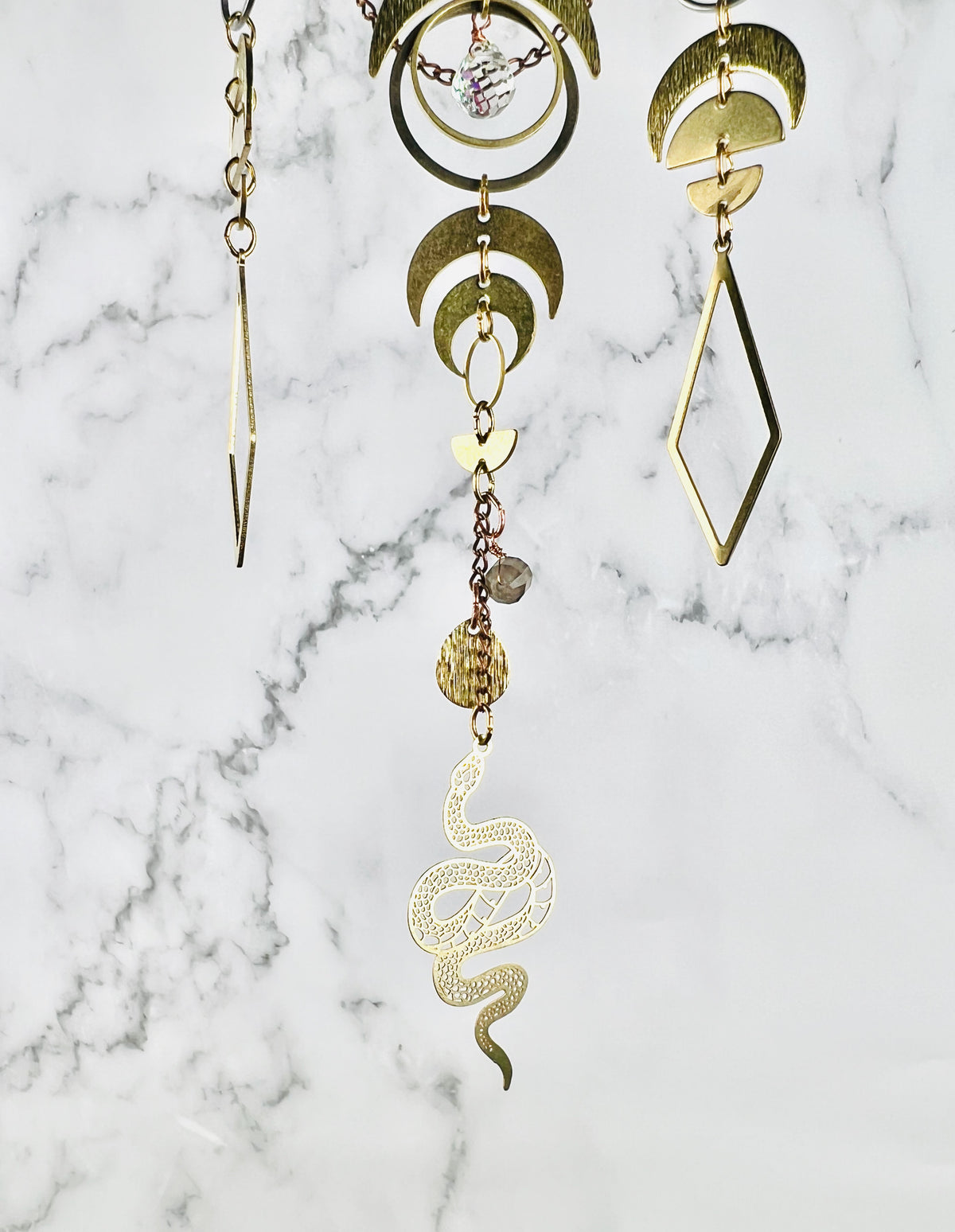 A close-up of three connected strands of gold-toned geometric and organic charm pieces hanging in front of a gray and white marble background, including crescent moons, half circles, diamonds, rings, and snake-shaped elements.