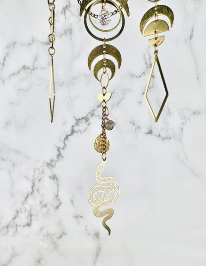 A close-up of three connected strands of gold-toned geometric and organic charm pieces hanging in front of a gray and white marble background, including crescent moons, half circles, diamonds, rings, and snake-shaped elements.