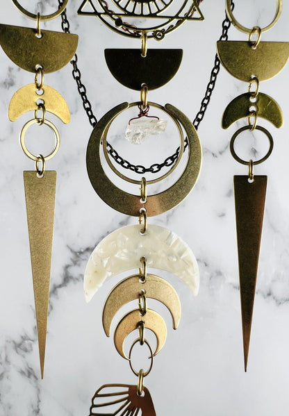 lower portion of wall art featuring brass-toned metal components including crescent moons, circles, and long triangular spikes.  A central white marbled acrylic crescent moon is suspended among the metal pieces, with a small clear citrine crystal accent all hanging in front of a marble wall.