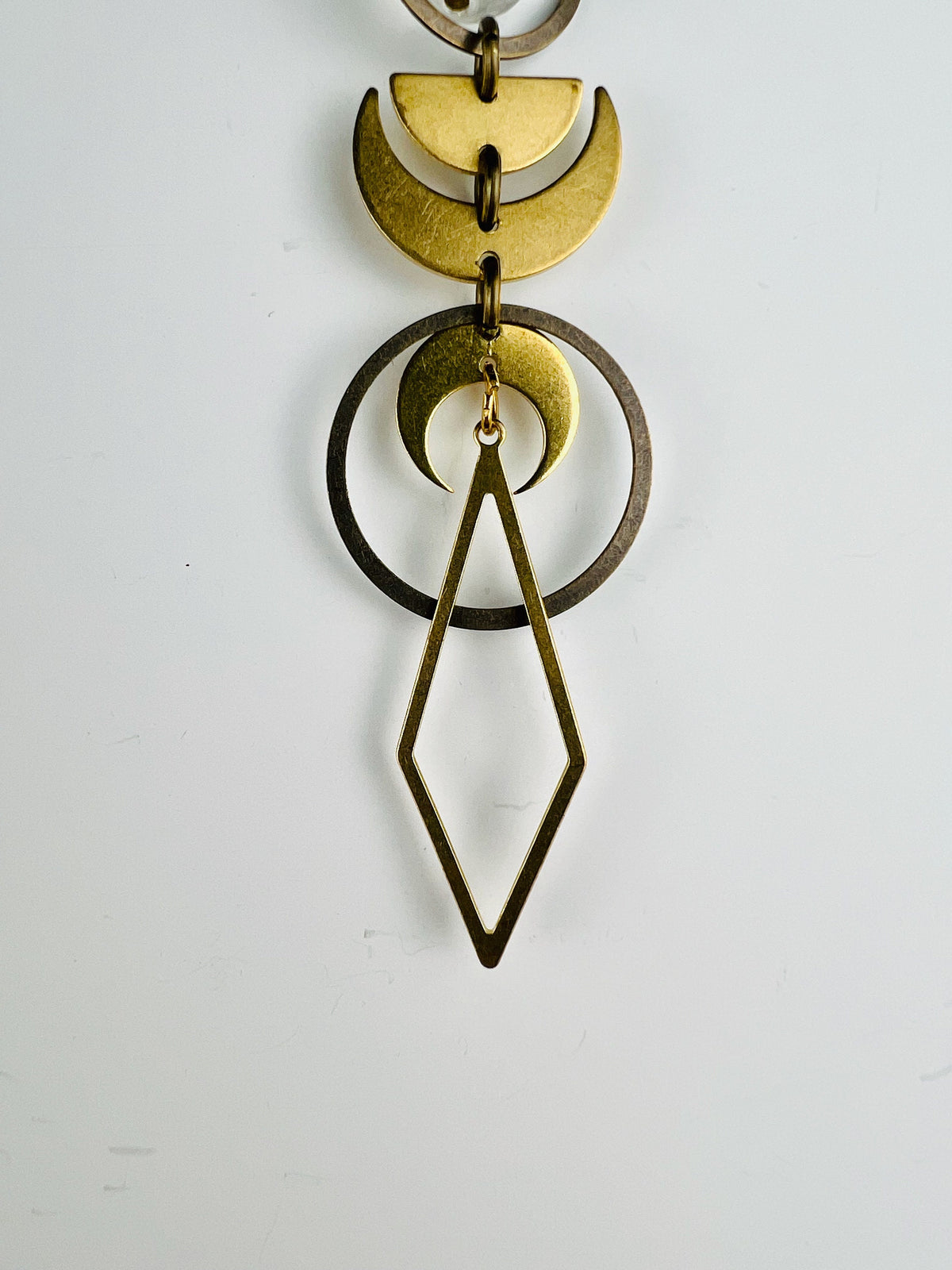 Gold colored moon face shapes such as crescent, moon, and half moon hang in a patterned with other shapes, such as bronze rings and a long diamond. They are connected by small gold colored rings and hang vertically in front of a light gray wall.
