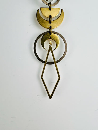 Gold colored moon face shapes such as crescent, moon, and half moon hang in a patterned with other shapes, such as bronze rings and a long diamond. They are connected by small gold colored rings and hang vertically in front of a light gray wall.