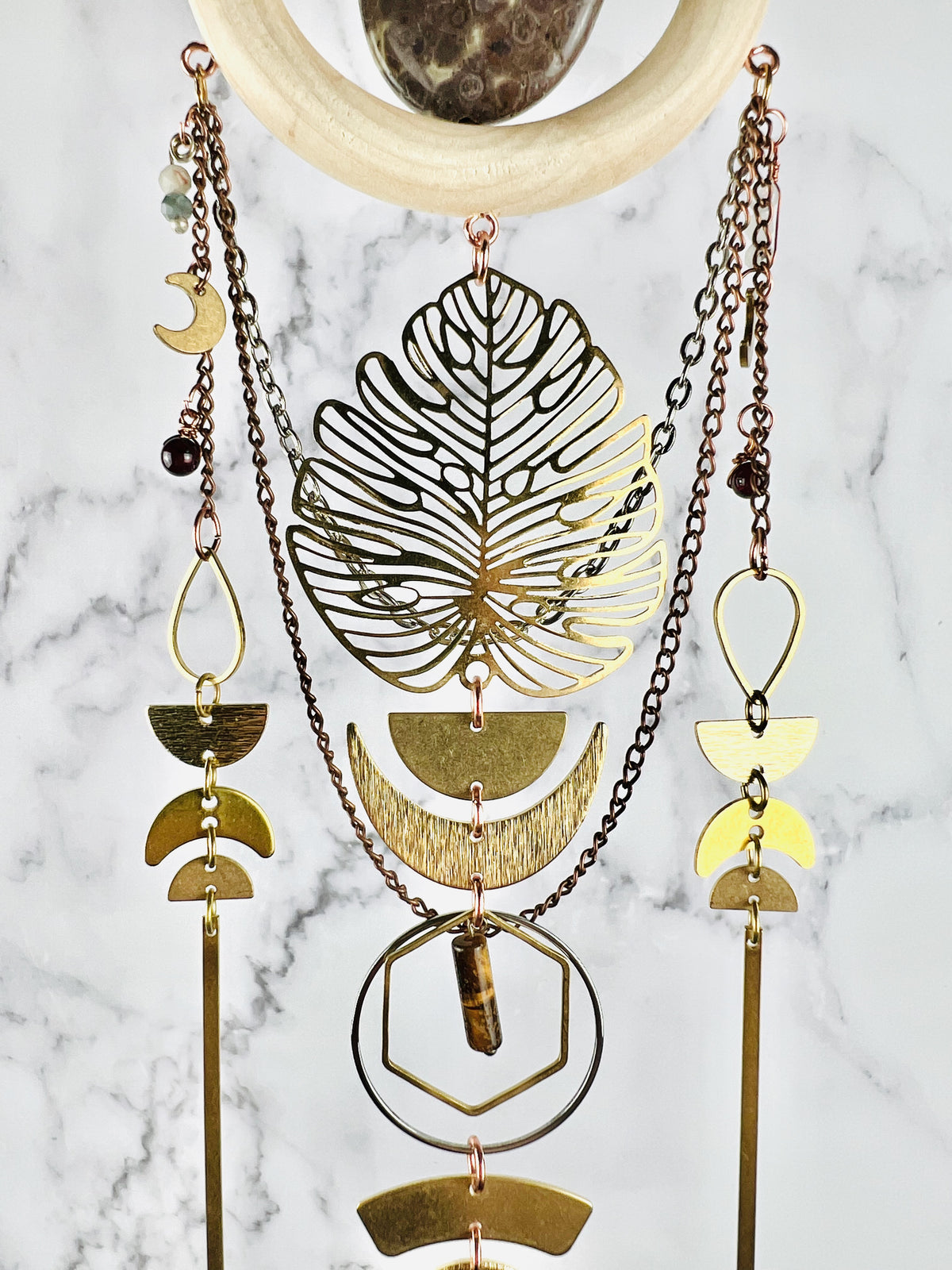 The bottom of a wood ring with a stone in the middle, hangs in front of a marble wall. Attached to the wood ring, hanging in a beautiful pattern are copper and silver colored chain, wire wrapped beads-including jasper, moonstone, garnet and tiger's eye. Crescent, moons, a large Monstera leaf, half circles, long bars, hexagons and rings also hang from this piece, all in gold and silver colored metal. 