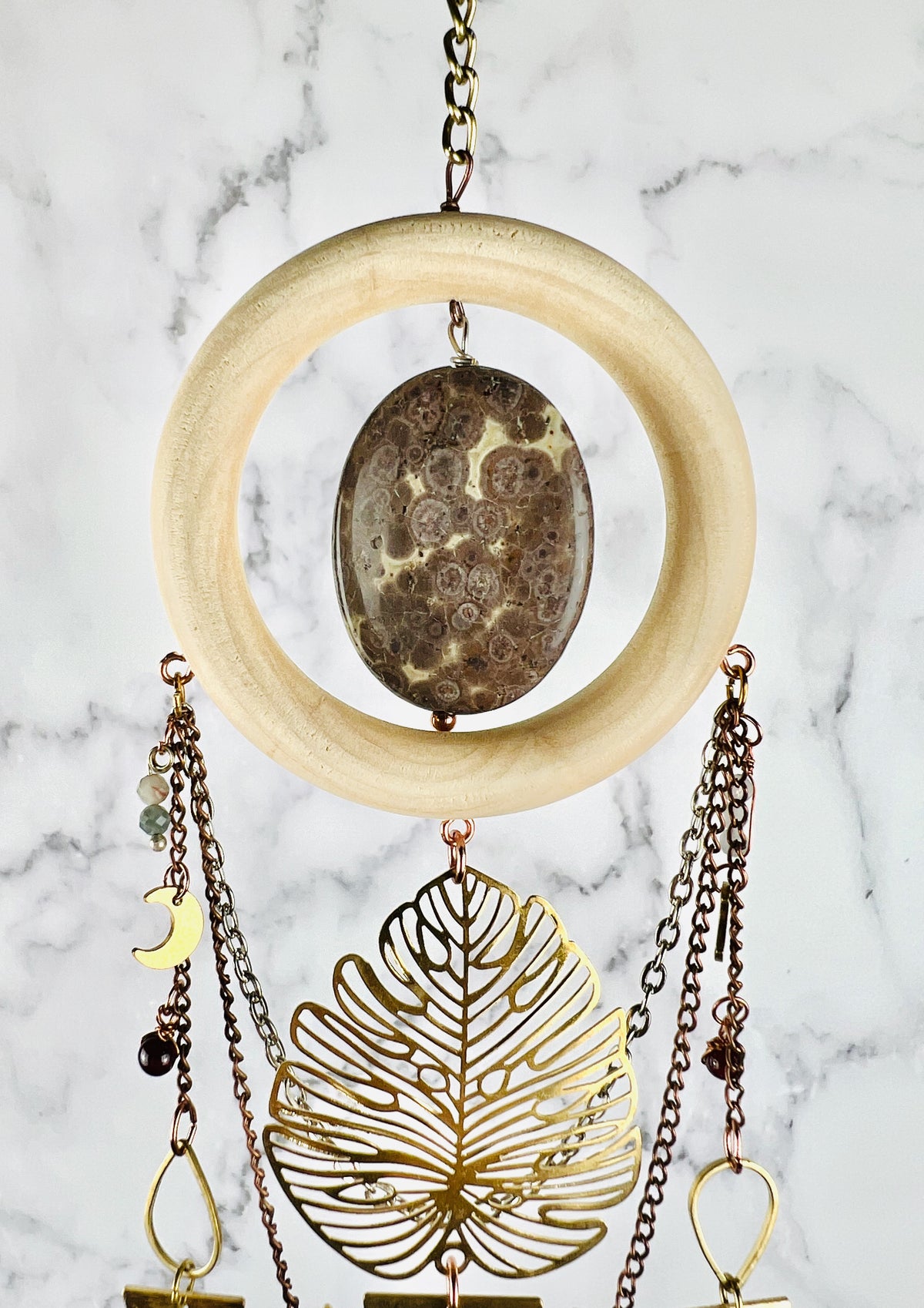 Hanging from a gold chain is a beautiful oval of Jasper surrounded by a light wood ring. Hanging from the lightwood ring are chains, a large Monstera leaf, wire wrapped beads, and various metal charms including crescent, moon and teardrop shapes. The piece hangs in front of a marble wall.