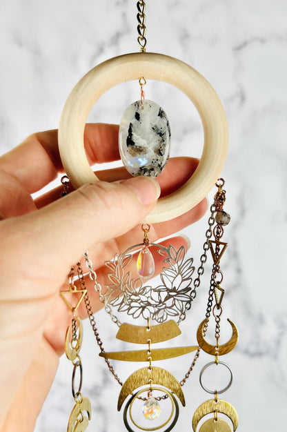 A hand holding a vertically hanging bohemian-style, crystal wall art featuring a light wood ring, a black-and-white oval moonstone, and multiple dangling gold and silver metal charms including: crescent moons, rings, triangles, a silver filigree flower wreath with prism at center and silver and copper colored chain. 