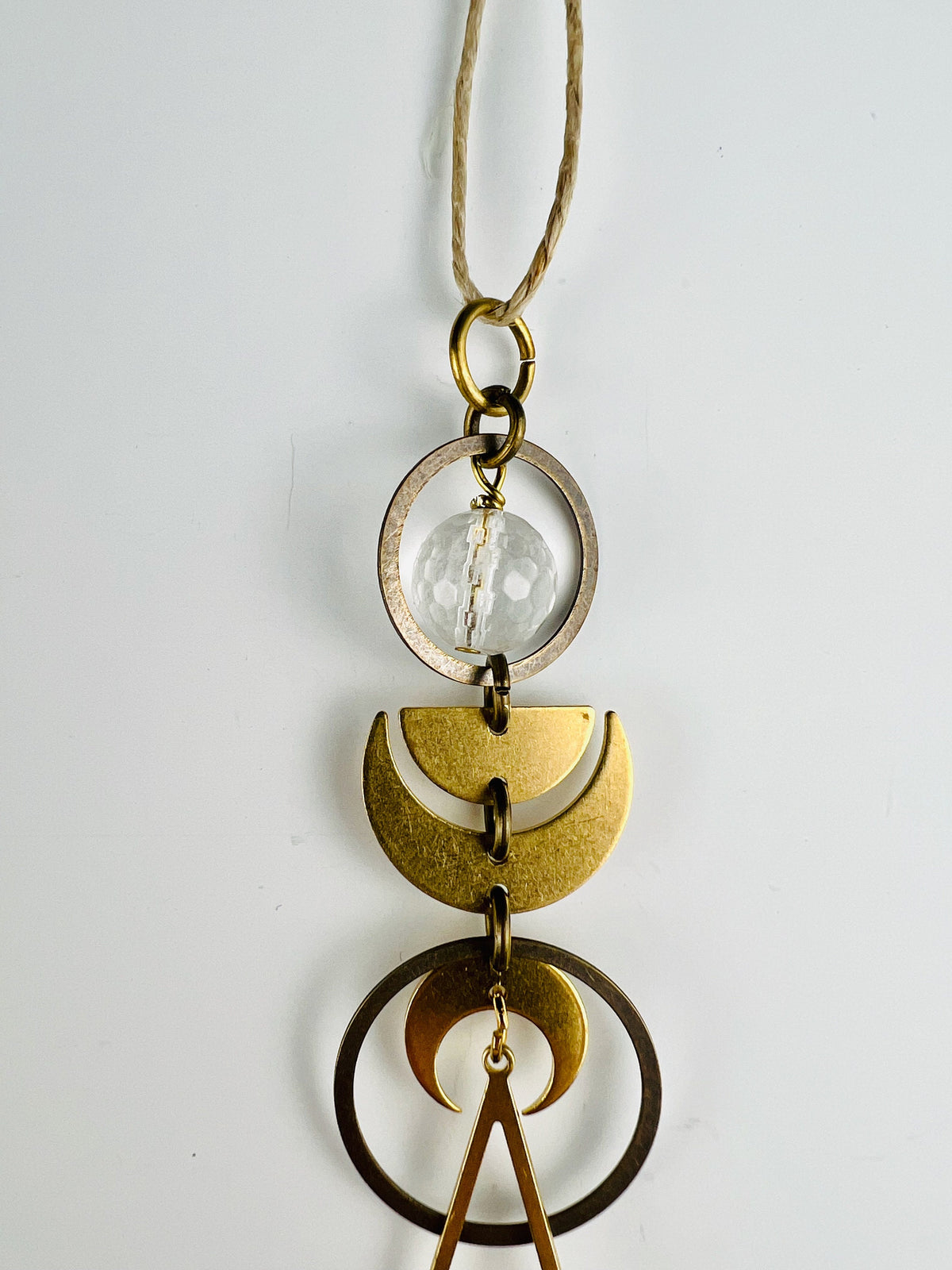 On a hemp string hangs a vertical metal rear view mirror Charm made with a faceted, clear quartz bead, bronze rings and gold colored metal moon phases with a pointed shape hanging from the bottom. The piece hangs in front of a light gray wall.