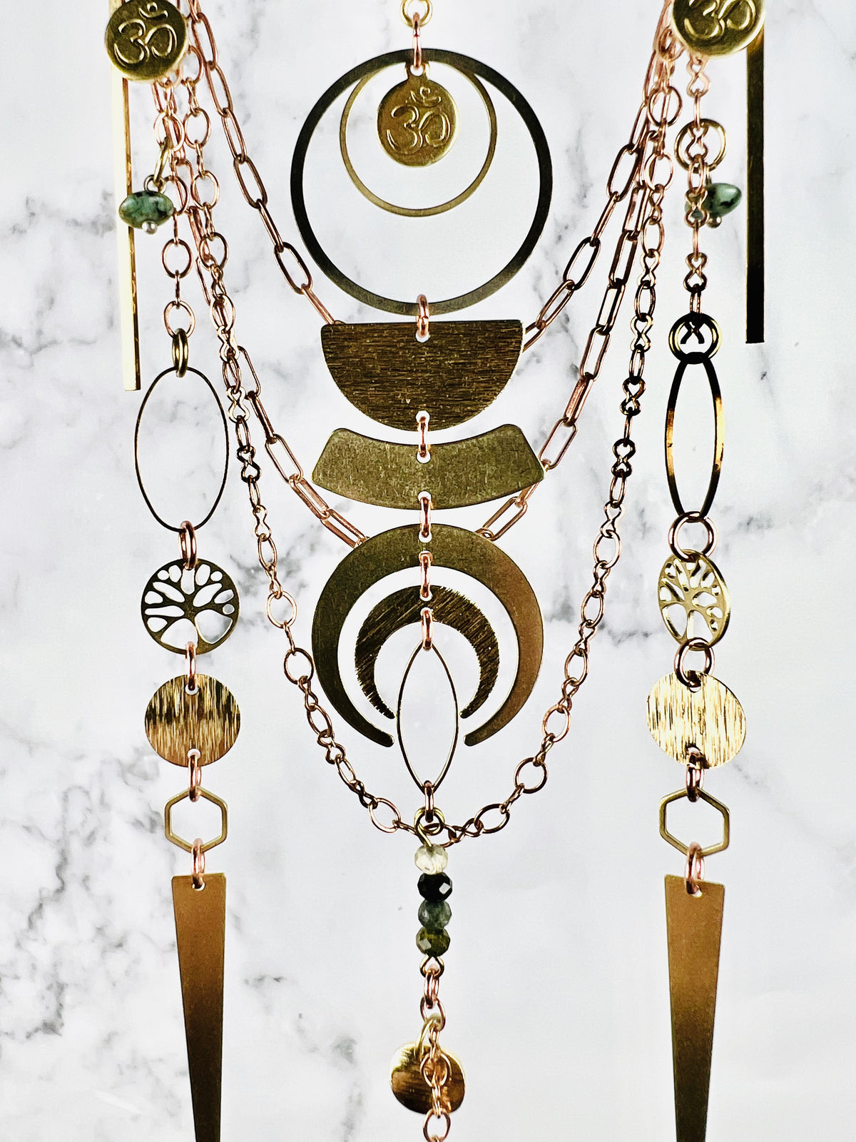 Copper colored chain with other various hanging metal pieces, including circles, tree of life, oh symbol, Rings, oval, half circles and Crescent Moon with wire wrapped beads at the bottom, hanging in front of a marble wall.