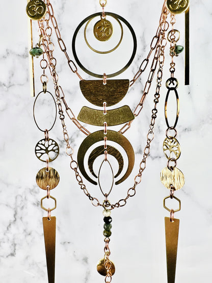 Copper colored chain with other various hanging metal pieces, including circles, tree of life, oh symbol, Rings, oval, half circles and Crescent Moon with wire wrapped beads at the bottom, hanging in front of a marble wall.