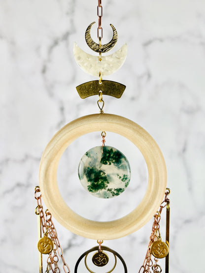 Hanging crystal wall art with copper color chain, brass crescent moons, acrylic crescent moon, Wood ring, and Om symbols, hanging in front of Of A marble wall.