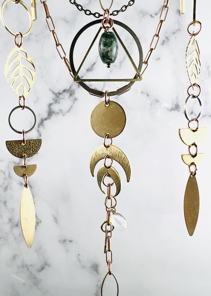 A multi-strand crystal wall hanging featuring brass and copper-toned metal components. The design includes geometric shapes like triangles, circles, leaves, crescent moons, half circles, and rings, accented with a green moss agate bead and a clear, translucent stone bead.