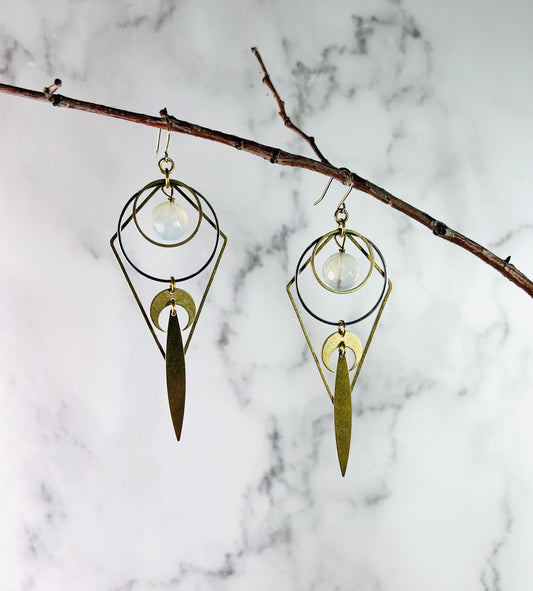 Handcrafted faceted Opalite crystal earrings with lightweight dangle styling and a reversible diamond-shaped detail for two looks in one. other components include steel and gold colored metal rings, crescent moons and a long pointed oval hanging at the ends. The pair of earrings hang on a thin branch in front of a marble wall.
