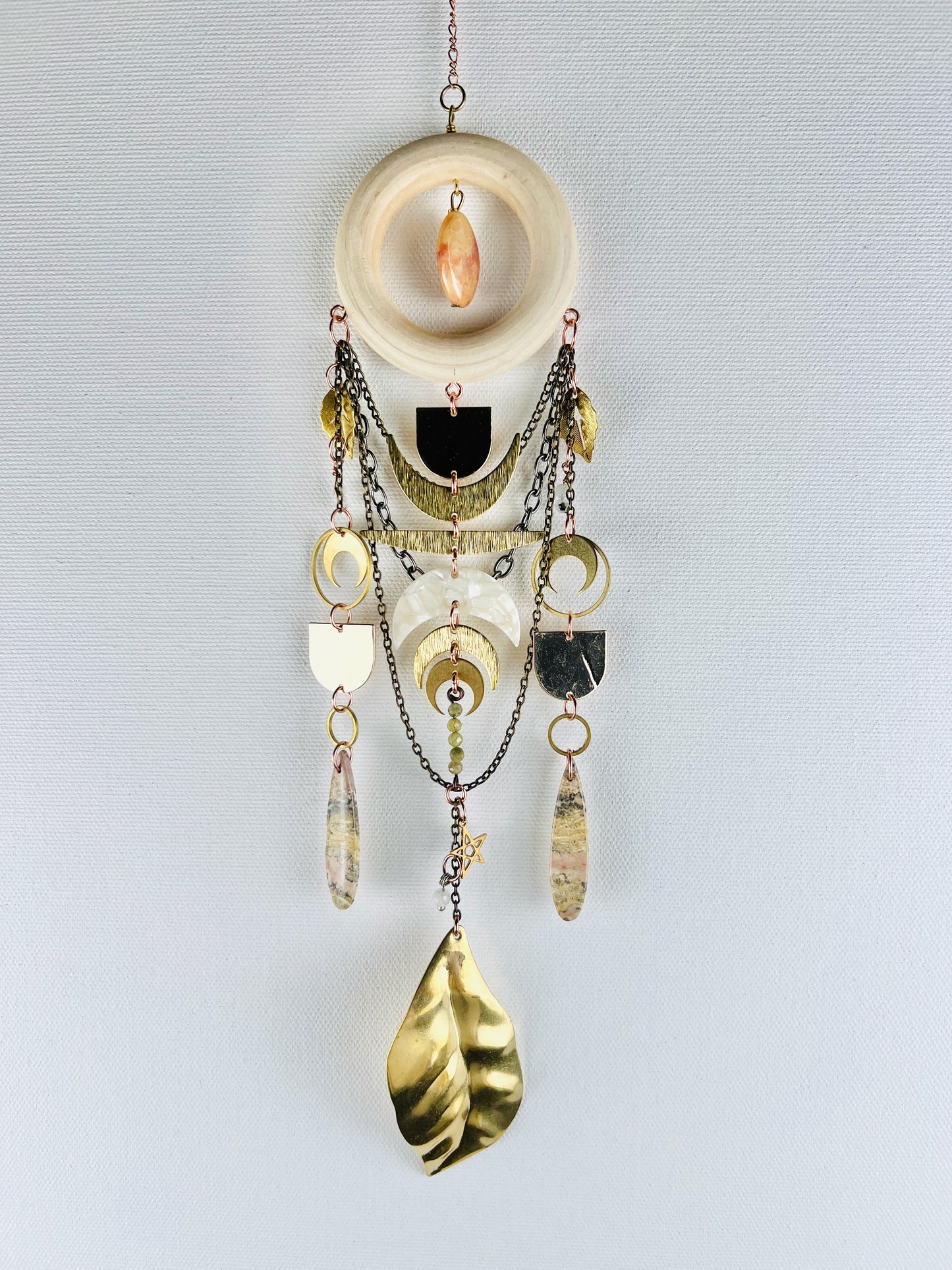 Decorative wall hanging with chain, gold leaves, rings, crescent moons, stars, jasper drop beads,  pink fossilized coral, and a wood ring on a light gray background