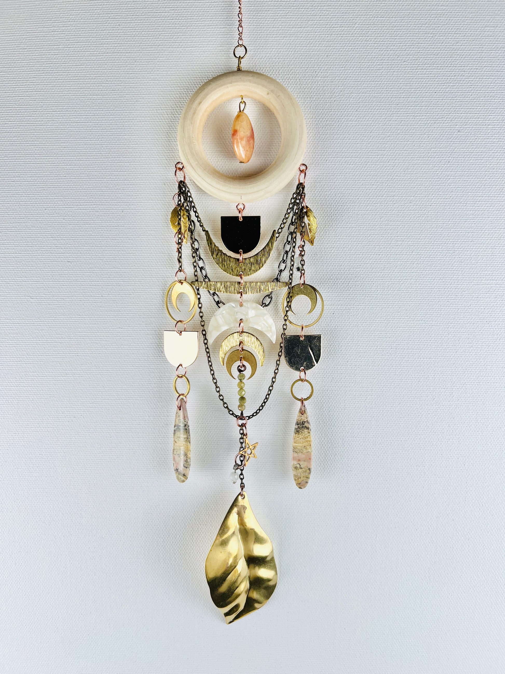 Decorative wall hanging with chain, gold leaves, rings, crescent moons, stars, jasper drop beads,  pink fossilized coral, and a wood ring on a light gray background