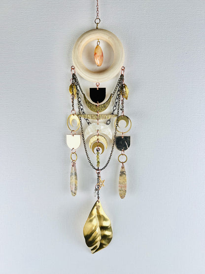 Decorative wall hanging with chain, gold leaves, rings, crescent moons, stars, jasper drop beads,  pink fossilized coral, and a wood ring on a light gray background