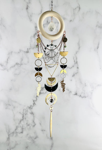 A piece of hanging crystal, wood and metal wall art, including elements, such as a cream colored wood ring with White drops shape stone in the middle, wire wrapped light blue beads, and gold colored metal geometric shapes, Fern leaves, moons, and a moth with moon phase above it all suspended in front of a gray and white marble wall.