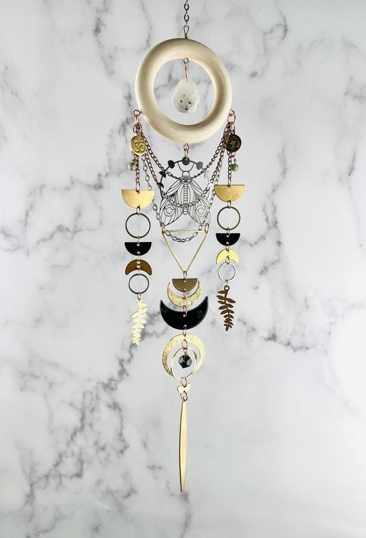 A piece of hanging crystal, wood and metal wall art, including elements, such as a cream colored wood ring with White drops shape stone in the middle, wire wrapped light blue beads, and gold colored metal geometric shapes, Fern leaves, moons, and a moth with moon phase above it all suspended in front of a gray and white marble wall.