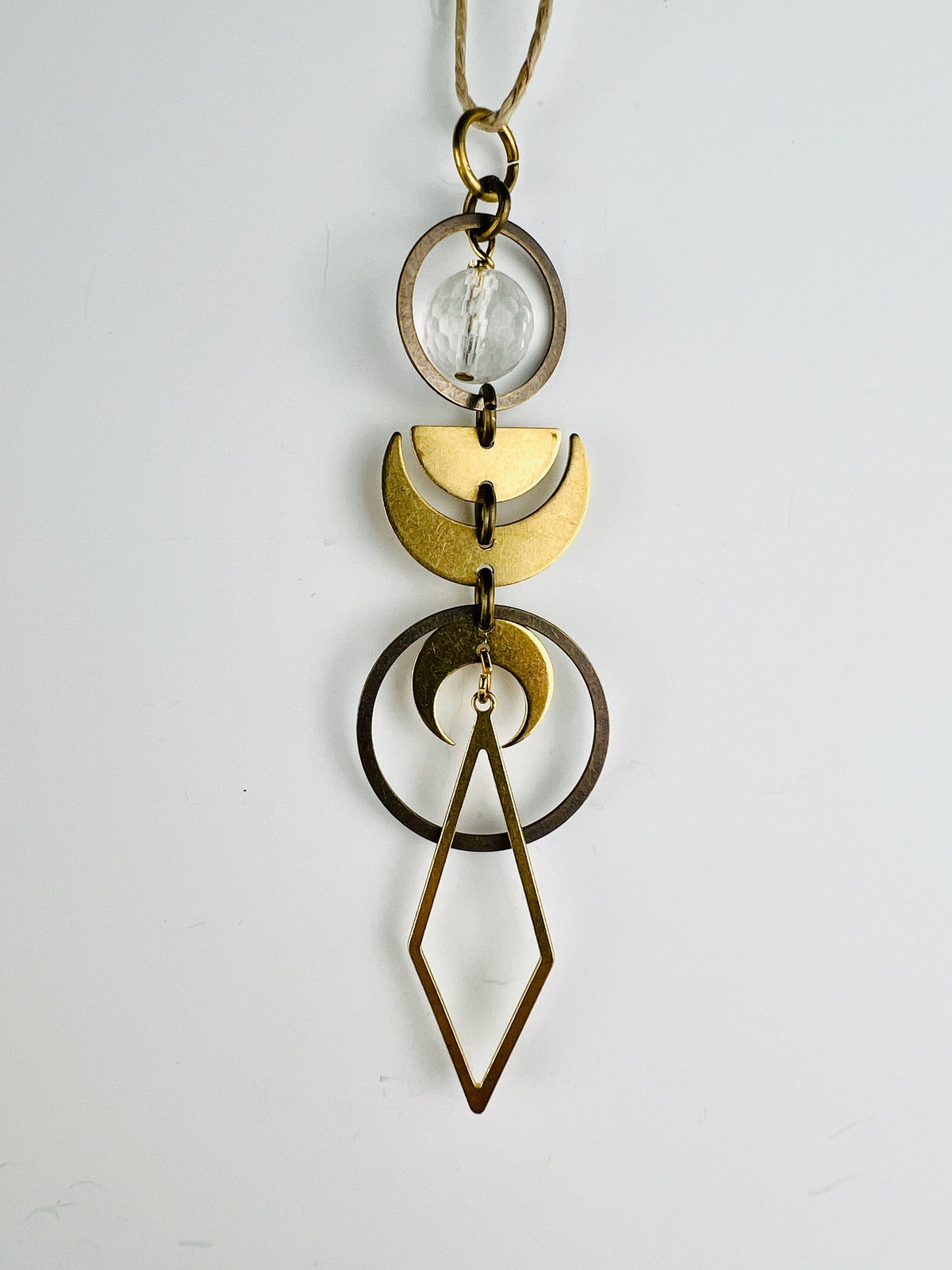 Clear Quartz rearview mirror charm measuring approximately 4 inches long and 1 inch wide, handcrafted as hanging crystal car decor. The Charm is a series of bronze rings with a faceted clear quartz bead, half moon and crescent moons hanging in a vertical pattern with a diamond shape at the end. The item hangs on a white wall from a hemp string.