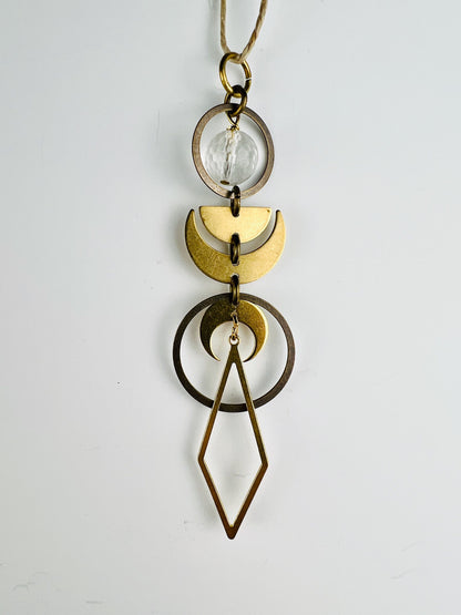 Clear Quartz rearview mirror charm measuring approximately 4 inches long and 1 inch wide, handcrafted as hanging crystal car decor. The Charm is a series of bronze rings with a faceted clear quartz bead, half moon and crescent moons hanging in a vertical pattern with a diamond shape at the end. The item hangs on a white wall from a hemp string.