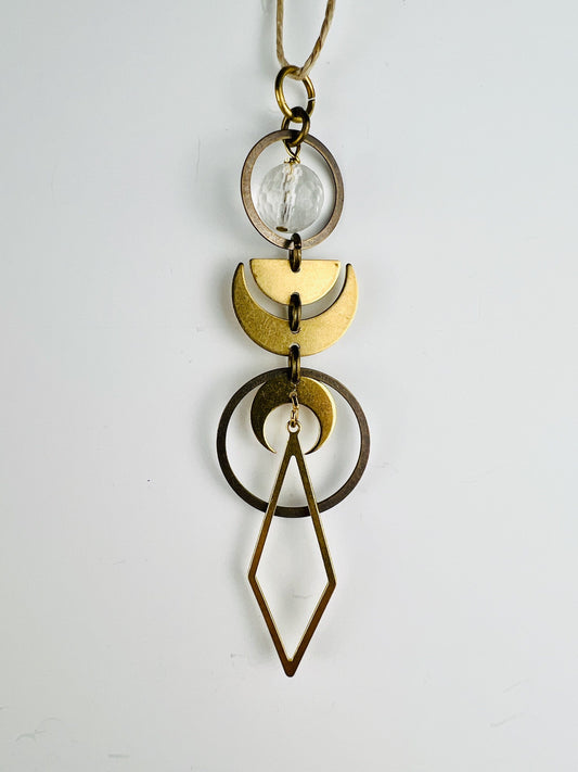 Clear Quartz rearview mirror charm measuring approximately 4 inches long and 1 inch wide, handcrafted as hanging crystal car decor. The Charm is a series of bronze rings with a faceted clear quartz bead, half moon and crescent moons hanging in a vertical pattern with a diamond shape at the end. The item hangs on a white wall from a hemp string.