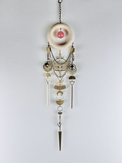 A crystal, metal and wood piece of wall art containing: a bronze chain attached to a cream colored wood ring, with a pink circular cherry quartz hanging in the center. Hanging vertically from it is a series of shapes, including: gold colored metal, moth and sword Charm, hexagons, bells, rings, crescent moons, half circles, triangles, and long bars along with wire wrapped beads and chain. All hangs on a light gray canvas wall.