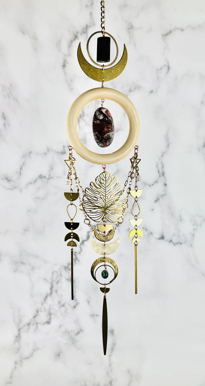 hypersthene, crazy lace agate, rose quartz and moss agate, dimensional crystal wall art hangs in front of a gray and white marble wall. Other elements on this vertically hanging art include: gold colored chain, rings, crescent, moons, an oval shaped red and grey stone in the middle of the cream colored wood ring, gold colored chain with prisms, triangles, half circles, teardrop shapes, and off-white acrylic, Moon, and long, slender gold bars hanging from the bottom of each three sections.