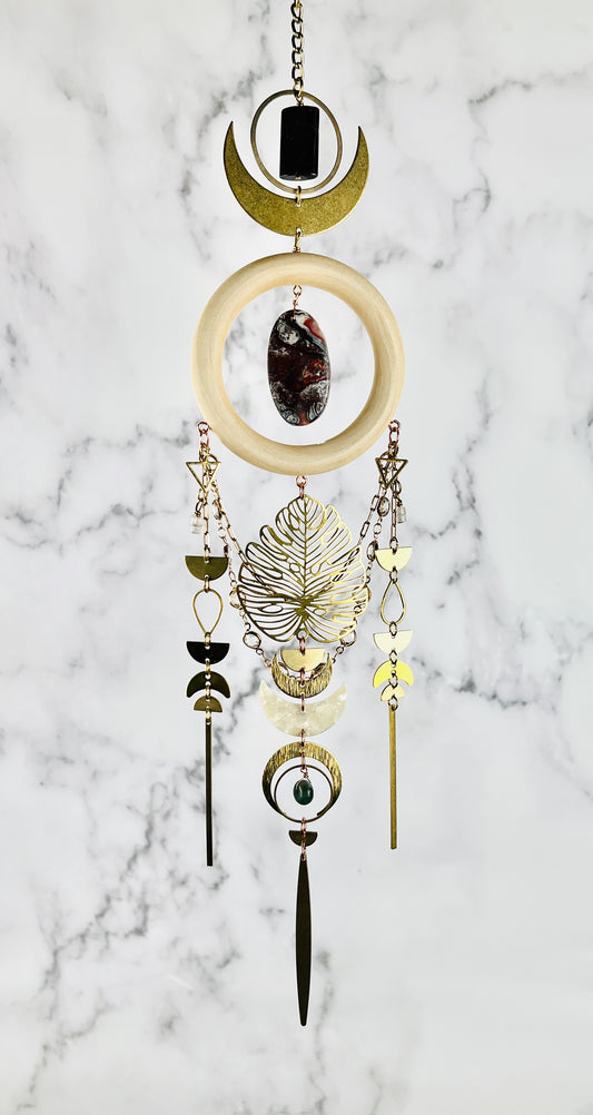 hypersthene, crazy lace agate, rose quartz and moss agate, dimensional crystal wall art hangs in front of a gray and white marble wall. Other elements on this vertically hanging art include: gold colored chain, rings, crescent, moons, an oval shaped red and grey stone in the middle of the cream colored wood ring, gold colored chain with prisms, triangles, half circles, teardrop shapes, and off-white acrylic, Moon, and long, slender gold bars hanging from the bottom of each three sections.