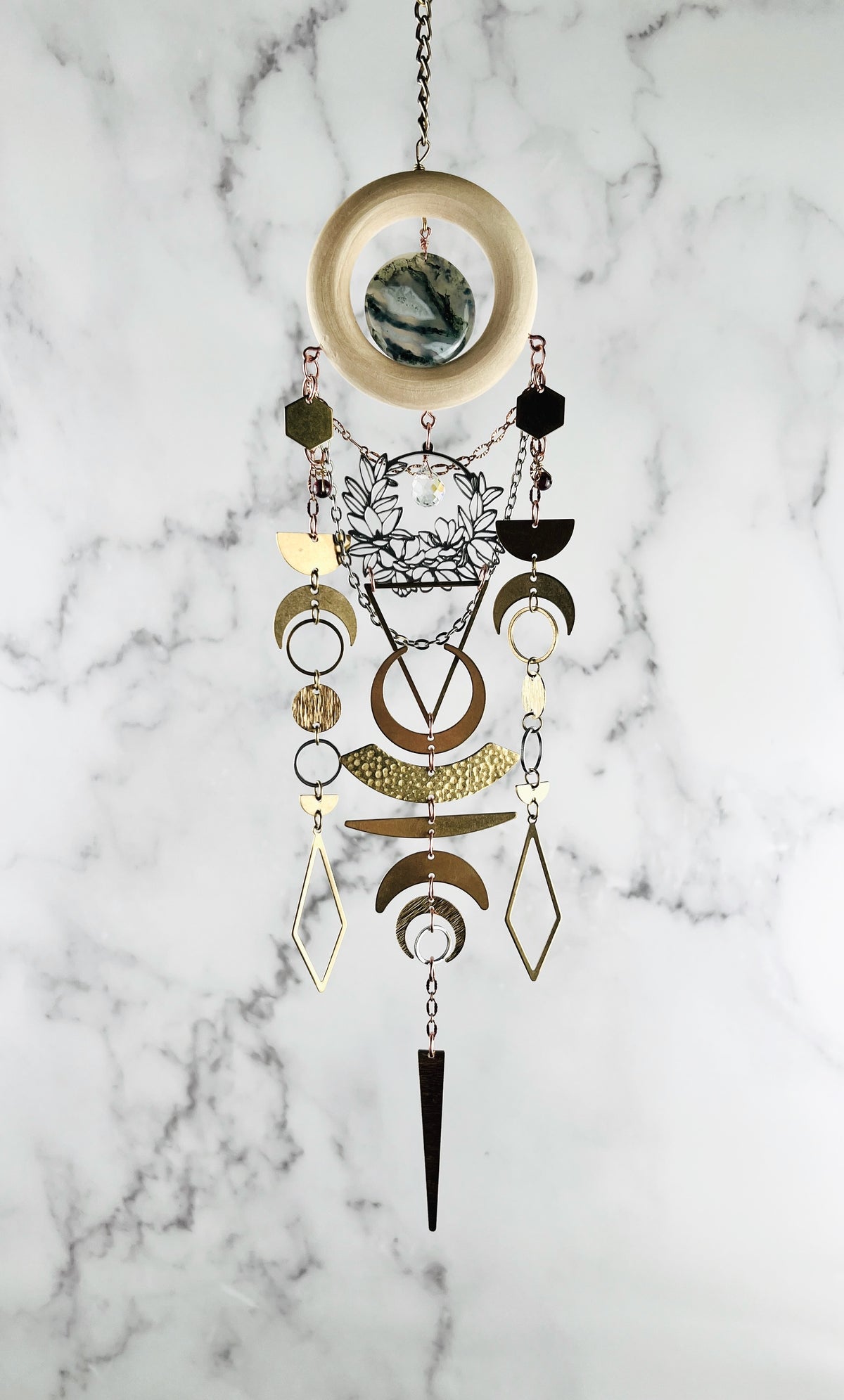 Decorative crystal hanging wall art with bronze chain, light wood ring, moss agate, gold colored metal geometric and nature shapes including: hexagons, diamonds, circles, triangles, half moons, crescent moons and silver flowers with prism, on a gray and white marble background. 