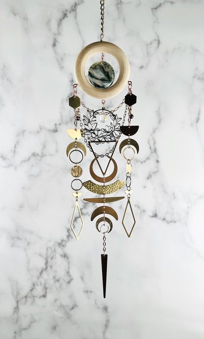 Decorative crystal hanging wall art with bronze chain, light wood ring, moss agate, gold colored metal geometric and nature shapes including: hexagons, diamonds, circles, triangles, half moons, crescent moons and silver flowers with prism, on a gray and white marble background. 
