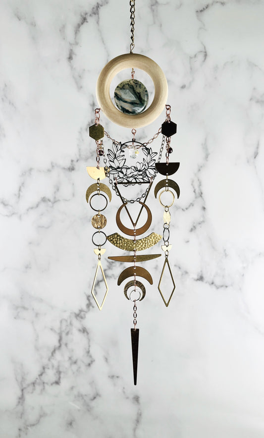 Decorative crystal hanging wall art with bronze chain, light wood ring, moss agate, gold colored metal geometric and nature shapes including: hexagons, diamonds, circles, triangles, half moons, crescent moons and silver flowers with prism, on a gray and white marble background. 