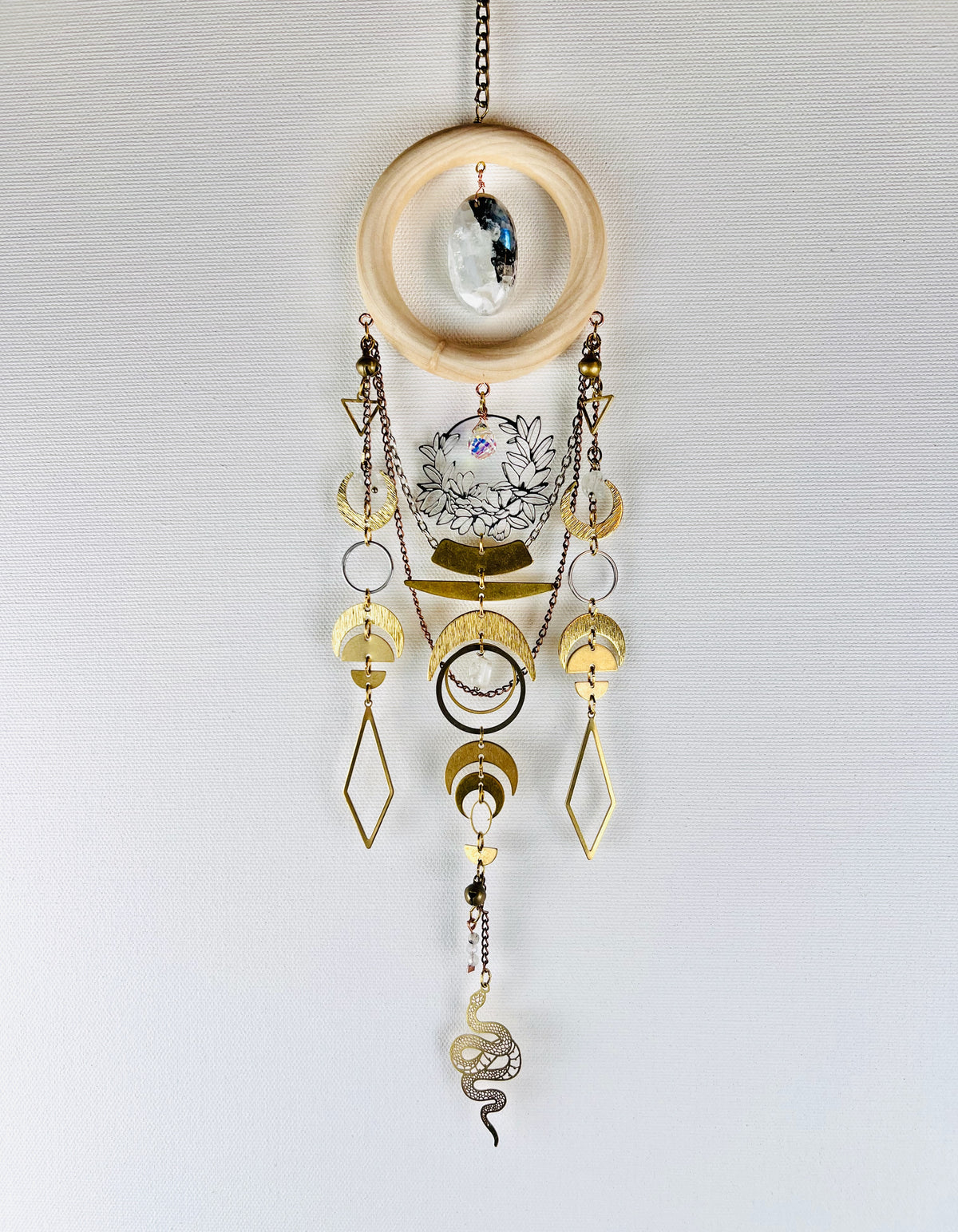 New beginnings, crystal wall art, hanging vertically in front of a light gray canvas with details, such as: a bronze chain, light wood ring with Center stone of oval of black and white moonstone, crystal, gold colored metal triangles, silver flower wreath charm with prism hanging in the middle, crescent, moons, silver rings, half circles, diamonds, wire wrapped beads, and a snake charm dangling at the bottom.