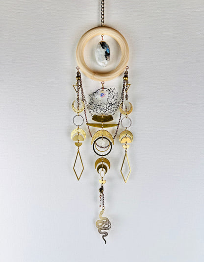 New beginnings, crystal wall art, hanging vertically in front of a light gray canvas with details, such as: a bronze chain, light wood ring with Center stone of oval of black and white moonstone, crystal, gold colored metal triangles, silver flower wreath charm with prism hanging in the middle, crescent, moons, silver rings, half circles, diamonds, wire wrapped beads, and a snake charm dangling at the bottom.