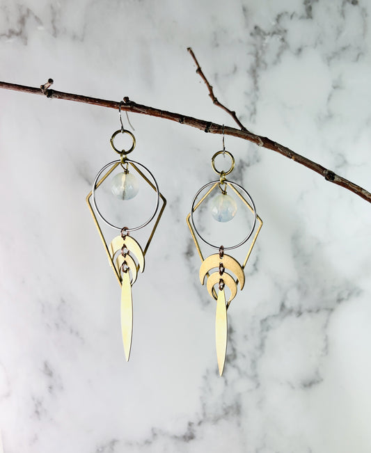 Pair of handcrafted faceted Opalite dangle earrings with reversible gold colored metal diamond-shaped and ring details, crescent moons and long pointed ovals at the bottom, measuring approximately 4 inches long and hanging on a thin branch in front of a marble wall.