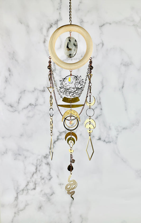 A bohemian-style piece of hanging crystal wall art against a gray and white marble background. The wind chime features a light wood ring, a central black and white moonstone crystal, a silver wreath of flowers with prism in center, gold-toned metal crescent moons and triangles, circles, and diamond charms, wire wrapped iolite beads and a small gold snake charm at the bottom.