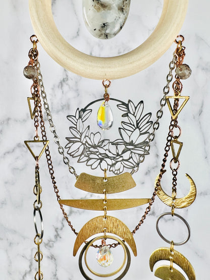 a close up photo of the upper middle portion of crystal wall, Art: a multiple natural material combined to form a piece of hanging dimensional art. It features vertical components attached together with tiny rings and an oval moonstone, surrounded by a light wood ring, wire wrapped iolite beads and various gold and silver colored metal charms such as: triangles, crescent moons, rings, a prism and silver and copper colored chain.