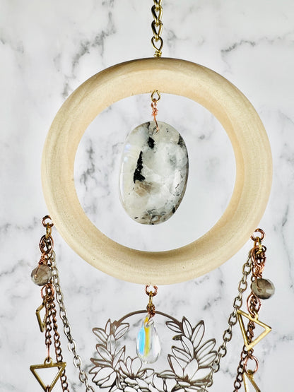 A bohemian-style hanging crystal wall art featuring a large natural wood ring with a polished white and black moonstone stone in the center, adorned with dangling chains, geometric gold colored metal charms, and a silver floral filigree piece against a gray and white marble background.