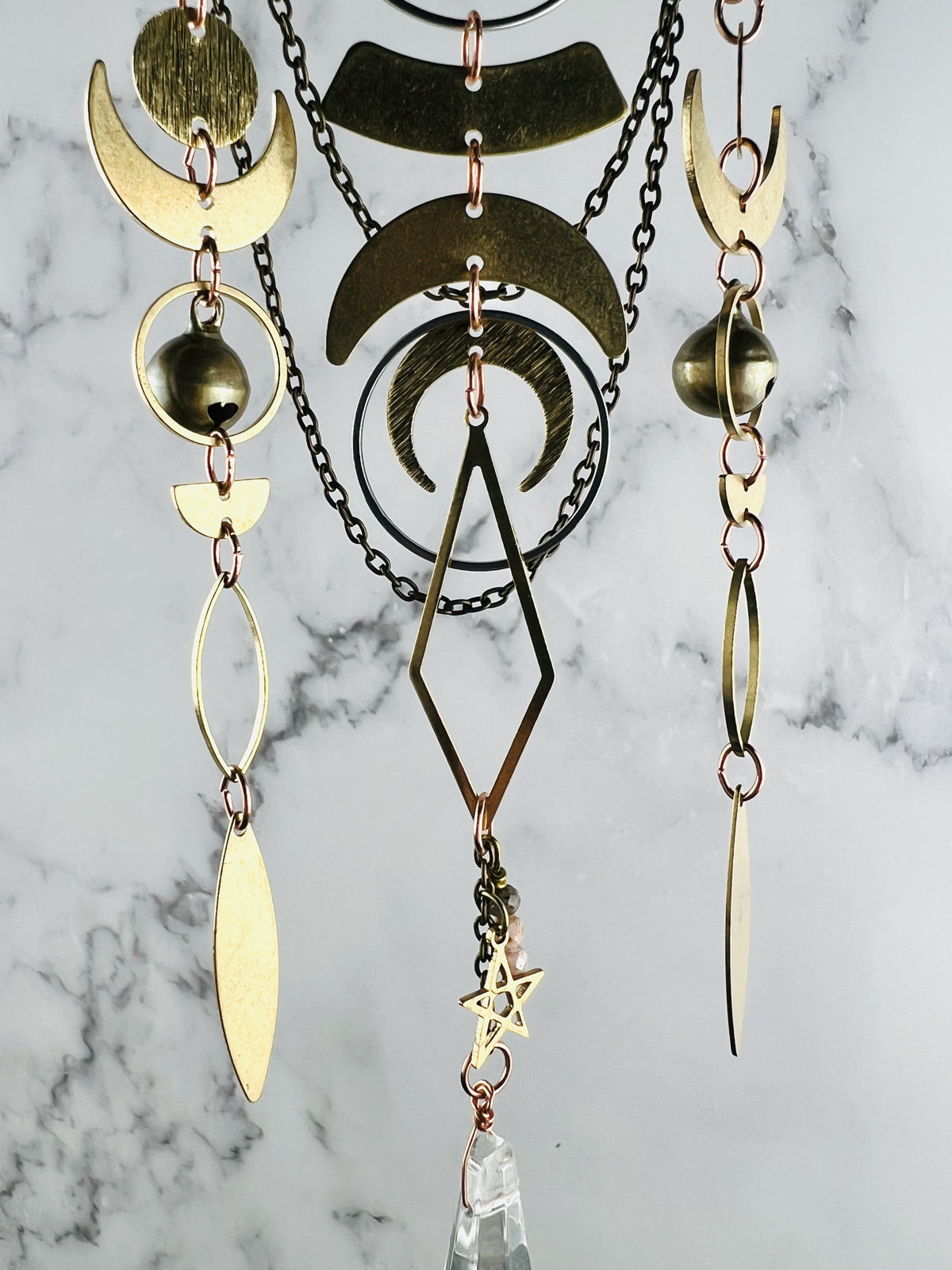 A gold and copper colored metal piece of hanging art with multiple crescent moon shapes, a diamond, and a star. The mobile has multiple chains and a clear crystal prism hanging from the bottom.