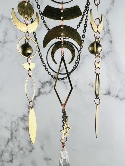 A gold and copper colored metal piece of hanging art with multiple crescent moon shapes, a diamond, and a star. The mobile has multiple chains and a clear crystal prism hanging from the bottom.