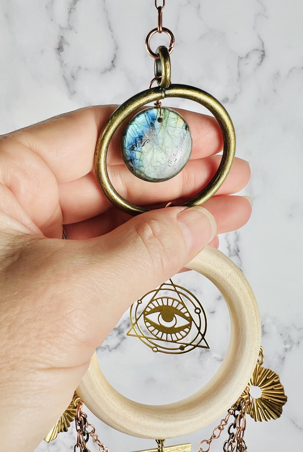 A hand gently grasps a labradorite, wire wrapped crystal at the top of a piece of wall art with light colored wood, a bronze ring, copper colored chain, an eye charm with triangle and rings around it and sunburst Charms. All hanging in front of a marble wall.