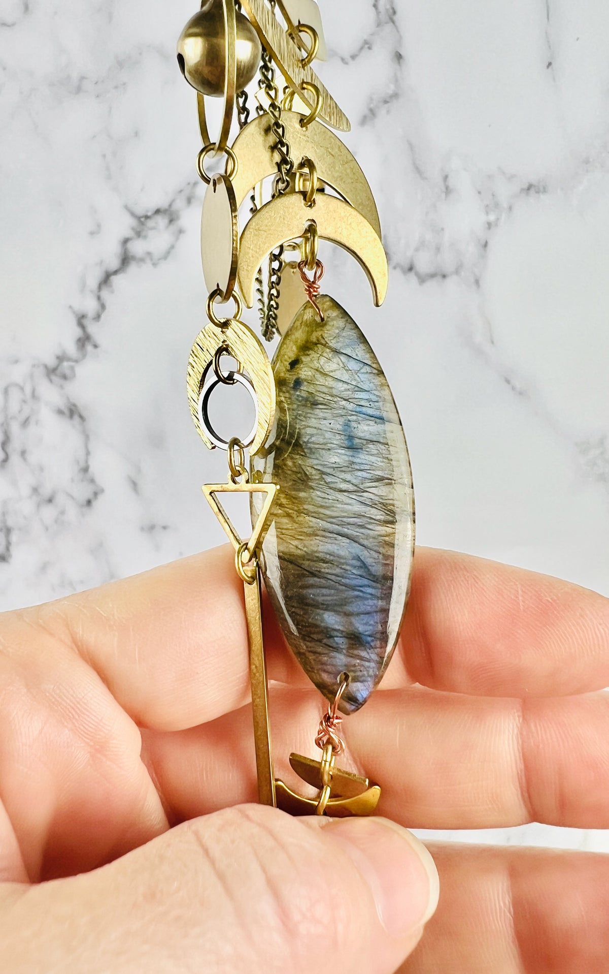 A hand grasps a marquise shaped piece of gray labradorite crystal from a piece of wall art. The peace also includes Brass crescent moons, a silver ring, a bell, rings, and oval, a triangle and a long gold bar.