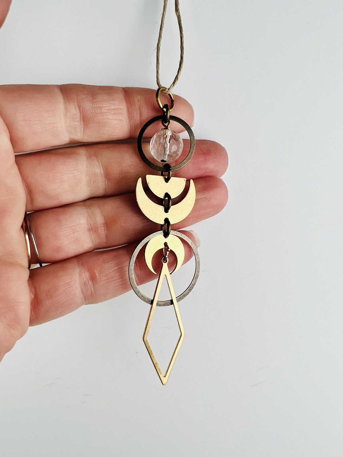 A hand with two rings holds a piece of vertically hanging gold colored metal art with components that are connected vertically with small rings. On this hanging crystal art are items, including: a round faceted clear quartz bead, bronze rings, crescent moons, and a long diamond shape hanging from the bottom. The charm hangs from a hemp string in front of a white wall.
