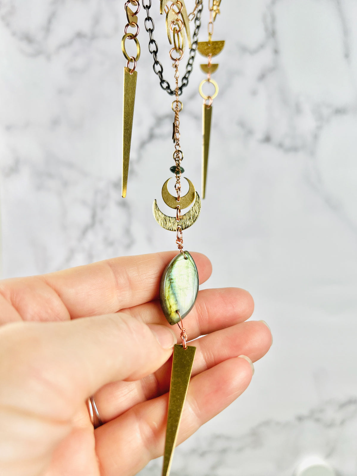 A hand gently lifts a labradorite bead on the bottom of a piece of crystal wall art with gold colored crescent moons, triangles, half circles, rings and chain in front of a marble wall.