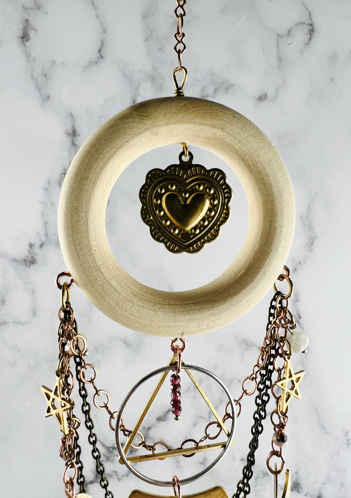 A light wood ring with a heart Charm in the middle, hangs from a chain in front of a marble wall. Also included on this piece of crystal wall art are dangling chains, stars, triangles, rings, and other wire wrapped beads.