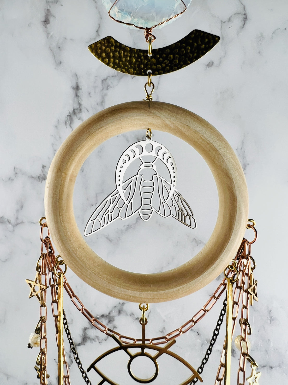 A decorative crystal wall hanging featuring a light wood ring, a silver bee charm with moon phases above it, a hammered brass arch, and a raw crystal top. Copper and brass chains dangle below with small star charms and a central eye-shaped metal piece.