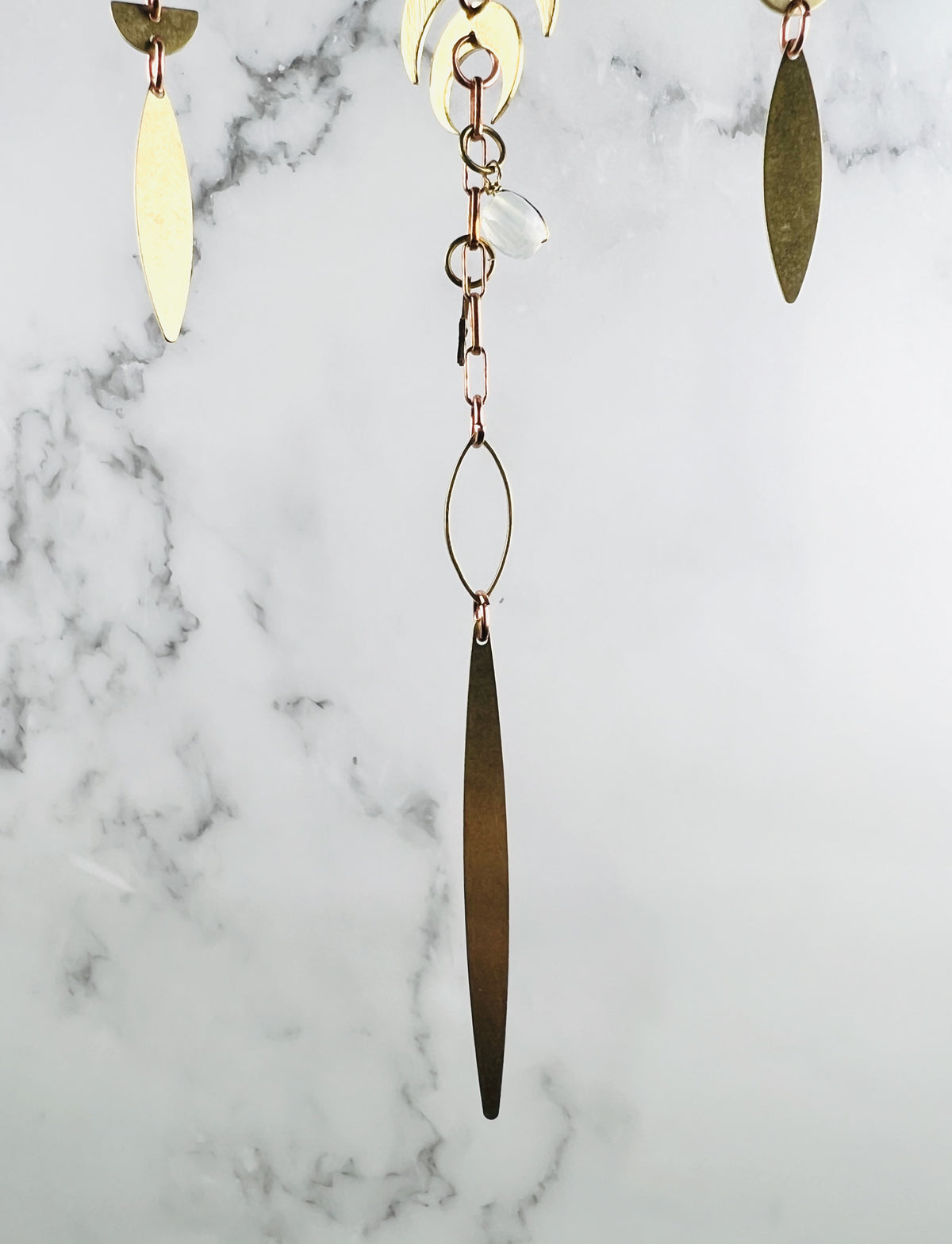 A hanging piece of dangling gold colored metal art, featuring multiple brass-toned components, including crescent moons, elongated leaf-like pendants, and thin oval wire frames, all connected by copper-colored metal chains and a single translucent white bead, in front of a marble wall.