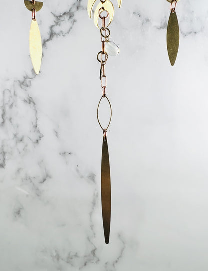 A hanging piece of dangling gold colored metal art, featuring multiple brass-toned components, including crescent moons, elongated leaf-like pendants, and thin oval wire frames, all connected by copper-colored metal chains and a single translucent white bead, in front of a marble wall.
