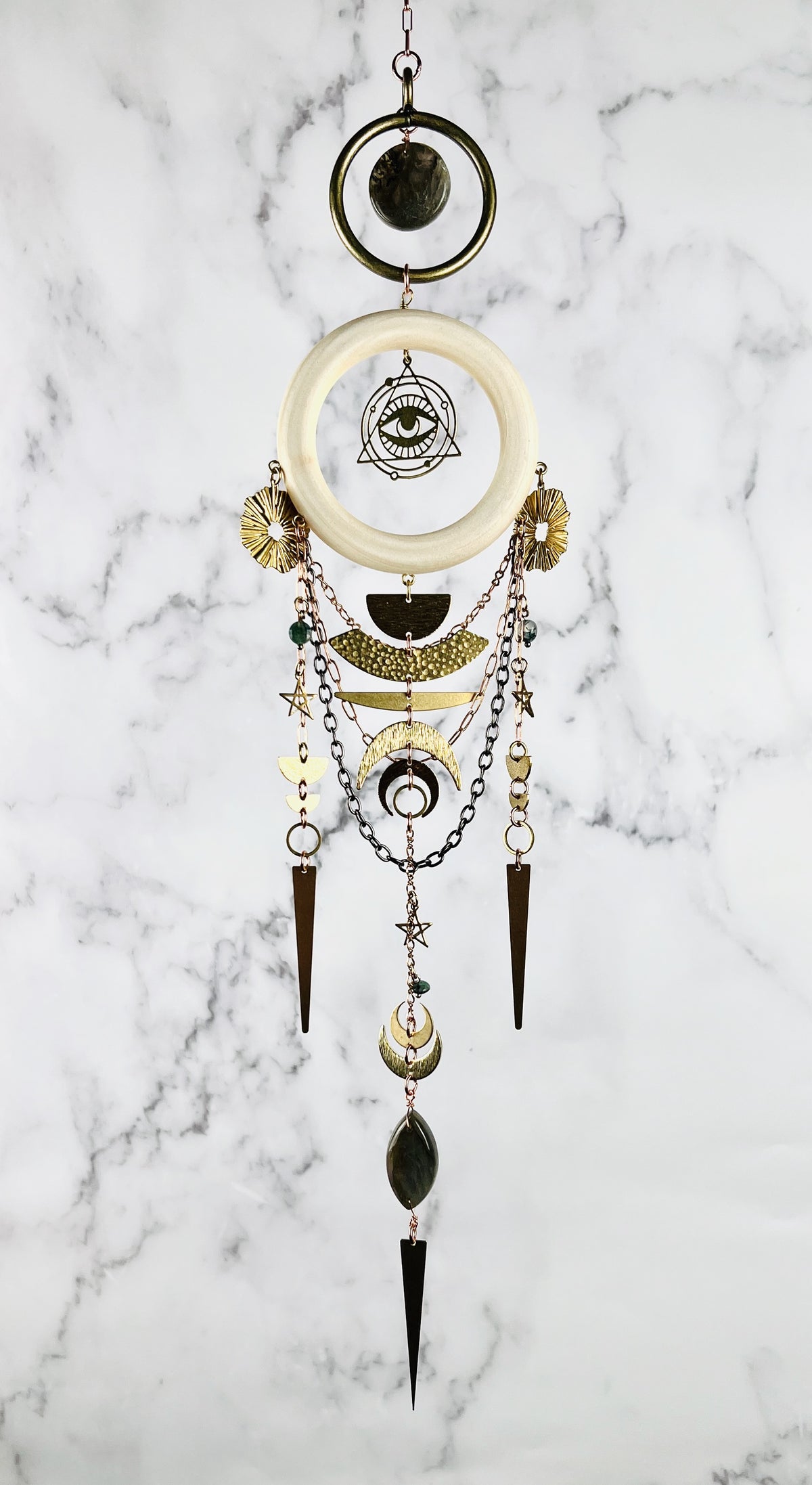 Hanging crystal wall art made with brass ring with natural Labradorite in the middle and below that a light wood ring hangs with an eye symbol surrounded by a triangle. Moss Agate stones arranged in a dimensional design, paired with gold colored metal, geometric shapes, and stars. The handcrafted piece measures approximately 16.5 inches tall and 4 inches wide and Hangs in front of a marble wall.