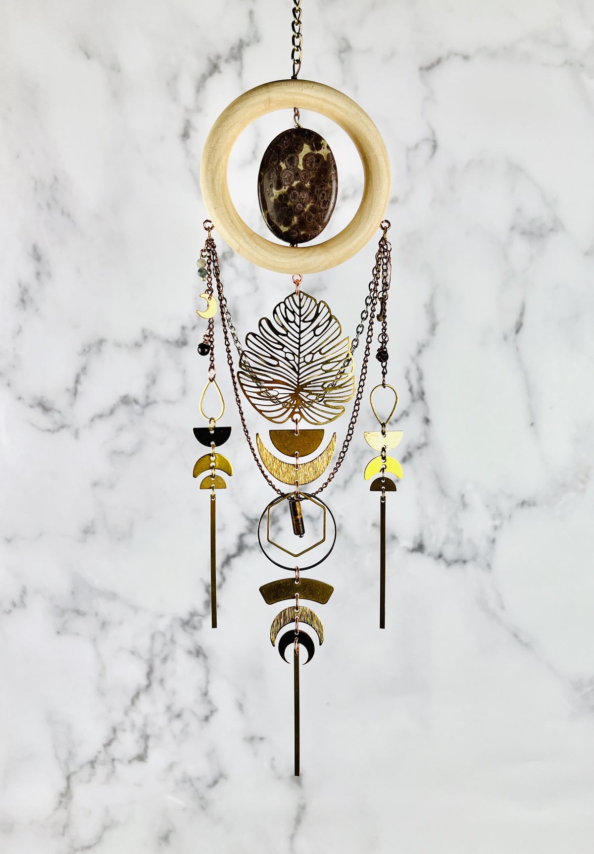 Handcrafted Jasper, Moonstone, Red Garnet and Tiger’s Eye crystal, wood and metal wall art. Features a wood ring with a mottled mauve and tan oval piece of Jasper in the center hanging from a chain. Chains also dangle from this Charm as well as wire wrapped beads and metal components, such as teardrop shapes, half circles, hexagons rings, crescent, moons, long gold bars and a large Monstera leaf.