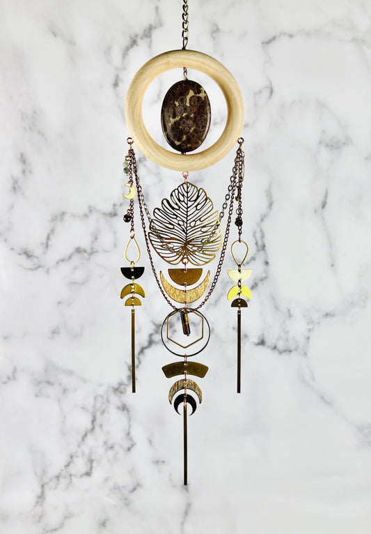 Handcrafted Jasper, Moonstone, Red Garnet and Tiger’s Eye crystal, wood and metal wall art. Features a wood ring with a mottled mauve and tan oval piece of Jasper in the center hanging from a chain. Chains also dangle from this Charm as well as wire wrapped beads and metal components, such as teardrop shapes, half circles, hexagons rings, crescent, moons, long gold bars and a large Monstera leaf.