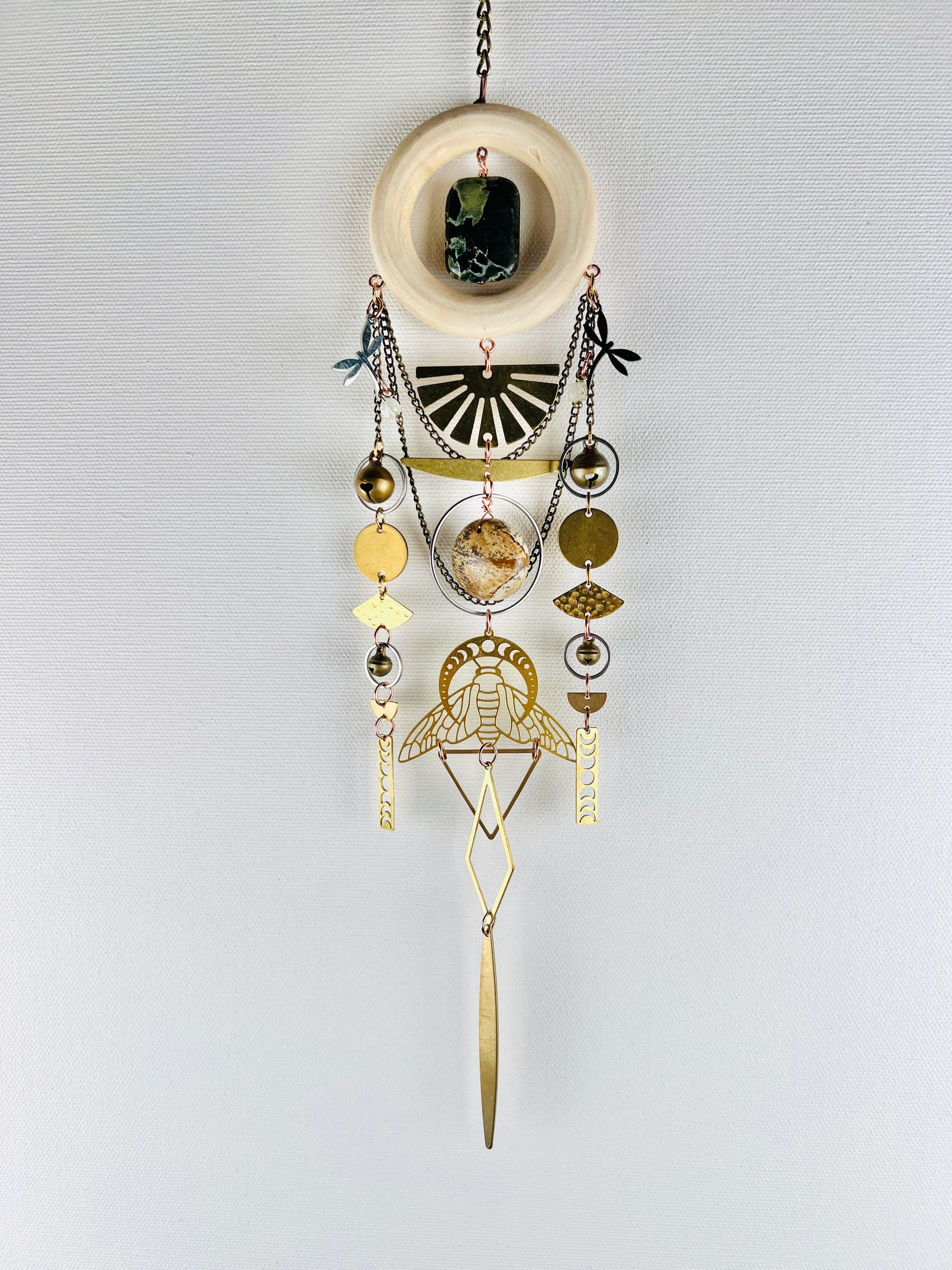 Decorative hanging crystal wall art with gold metal circles, moon phase, bells, triangles, and chain. also has silver dragon flies, and rings with a wood ring at the top; elements on a light gray background